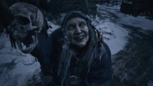 Mother Miranda from Resident Evil Village wearing a dark hood and eerie grin, surrounded by snow and skulls.